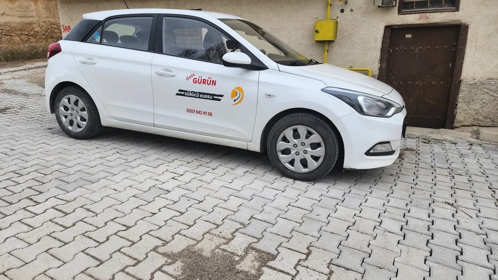 Yan kapısında Özel Gürün Sürücü Kursu logosu ve telefon numarası olan beyaz hatchback eğitim aracı, kaldırım taşlı sokakta