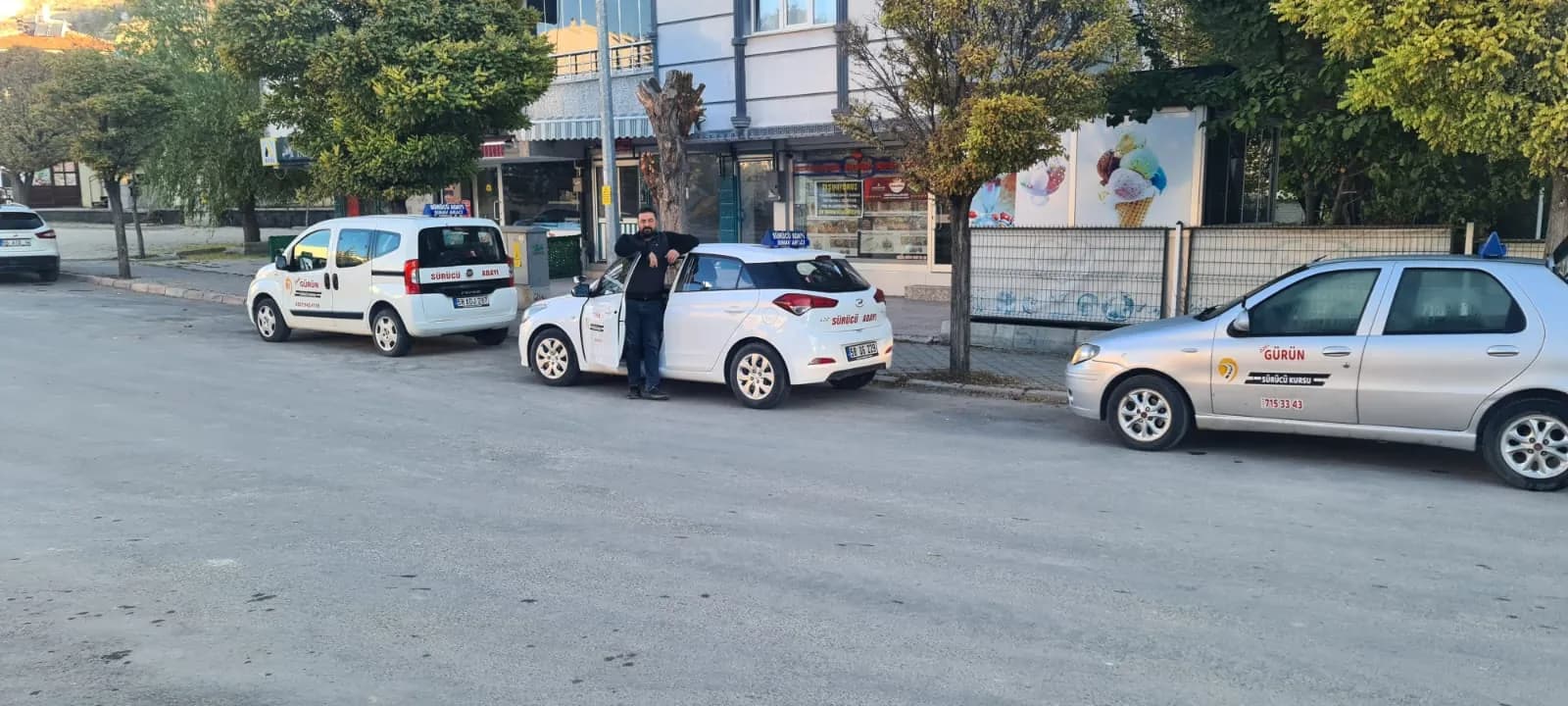 Sokak kenarında park halindeki üç Gürün Sürücü Kursu eğitim aracı; ortadaki aracın açık kapısında bir erkek duruyor