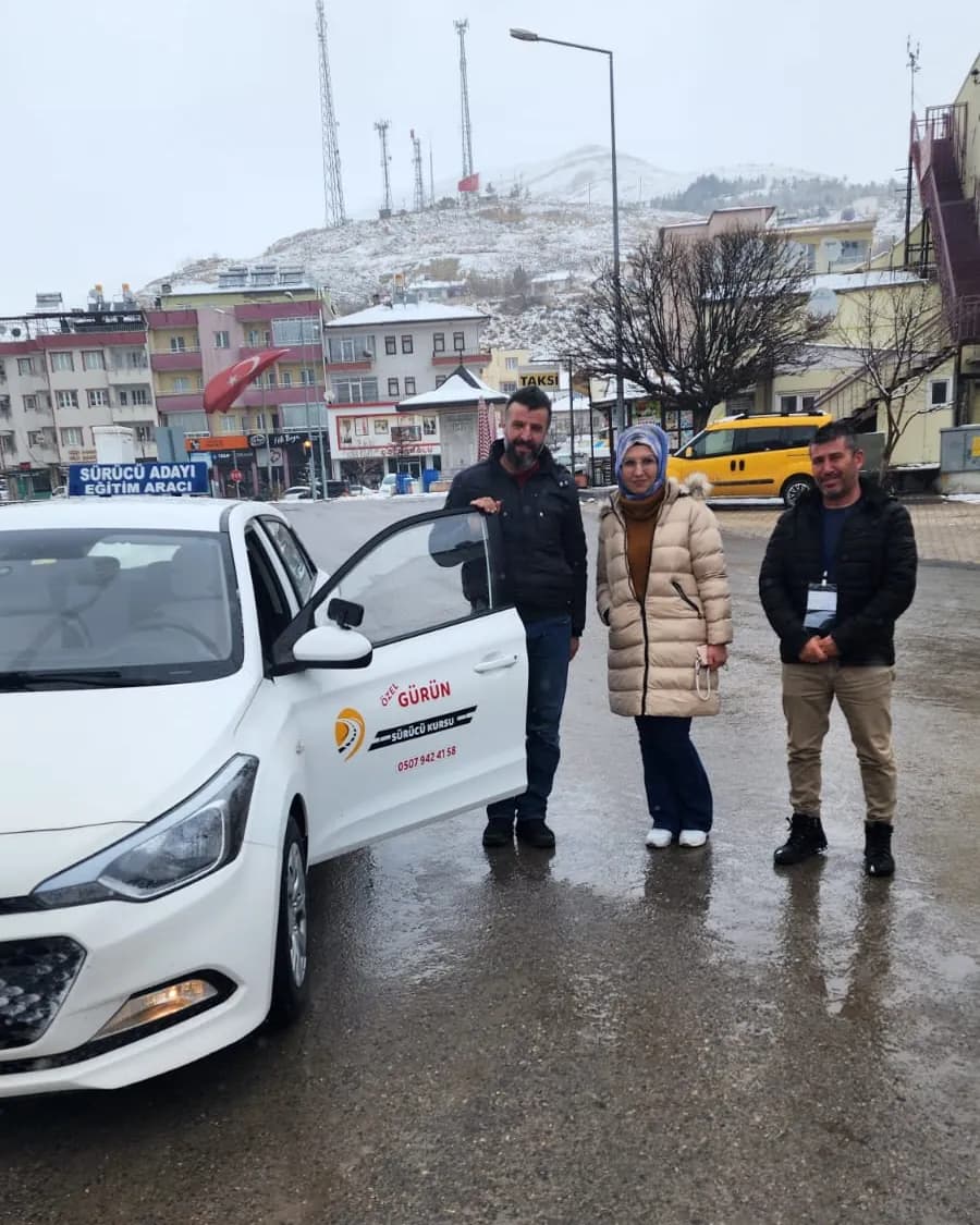 Karlı günde ıslak asfaltta beyaz eğitim aracının açık kapısı yanında duran iki erkek ve bir kadın; arkada tepe ve bayrak