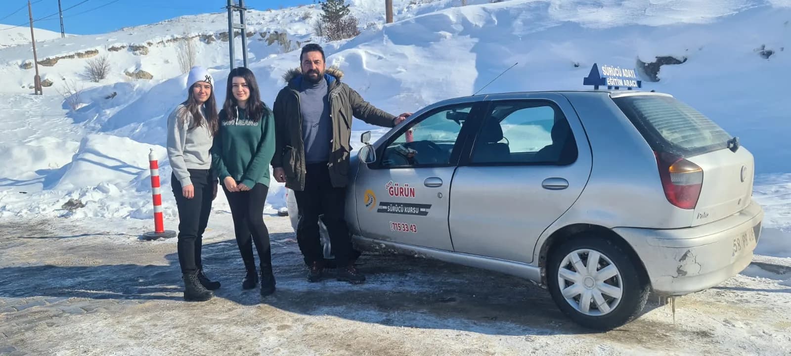 Karla kaplı yamacın önünde gümüş renkli eğitim aracının yanında poz veren iki genç kadın ve bir erkek
