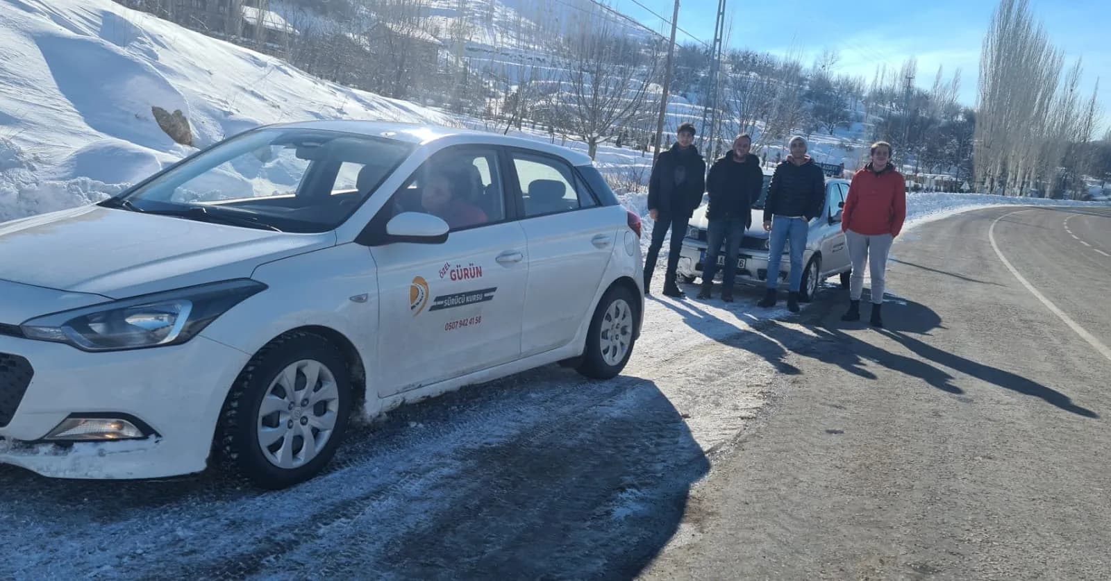 Karla kaplı yol kenarında park etmiş beyaz eğitim aracı; içinde bir kişi görünüyor, arkasında dört genç ayakta duruyor