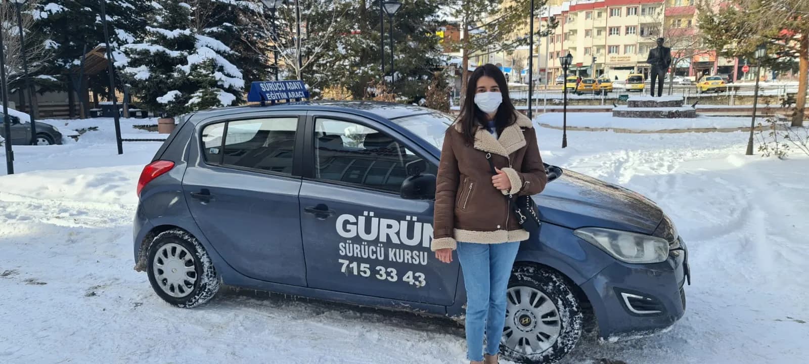 Karla kaplı parkta mavi-gri eğitim aracının önünde duran maskeli genç kadın; kapıda Gürün Sürücü Kursu yazısı var