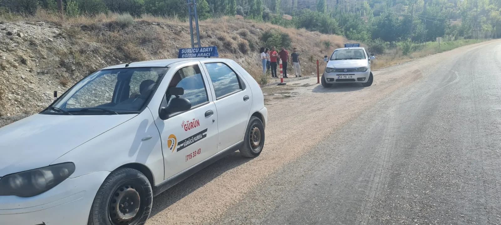 Kırsal yol kenarında iki Gürün Sürücü Kursu eğitim aracı; arka planda ayakta bekleyen küçük bir grup ve turuncu dubalar