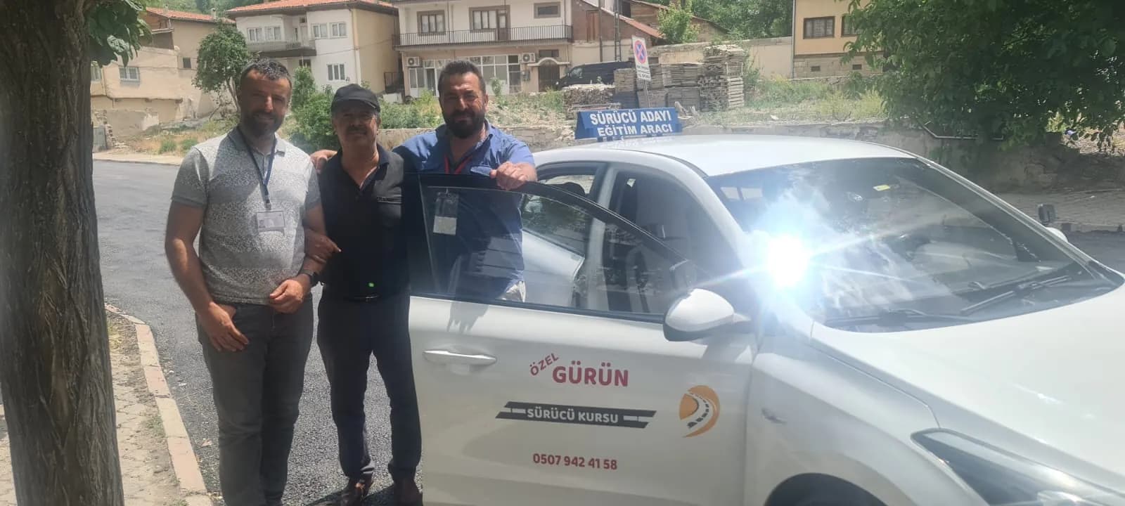 Gürün Sürücü Kursu eğitim aracının açık kapısı yanında yan yana poz veren üç erkek; ikisi yaka kartı takıyor