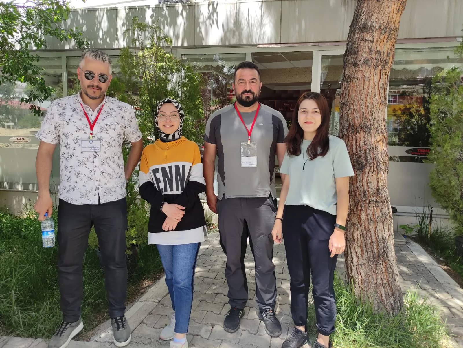 Ağacın yanında poz veren iki erkek ve iki kadın; erkeklerin boynunda kırmızı askılı yaka kartı var