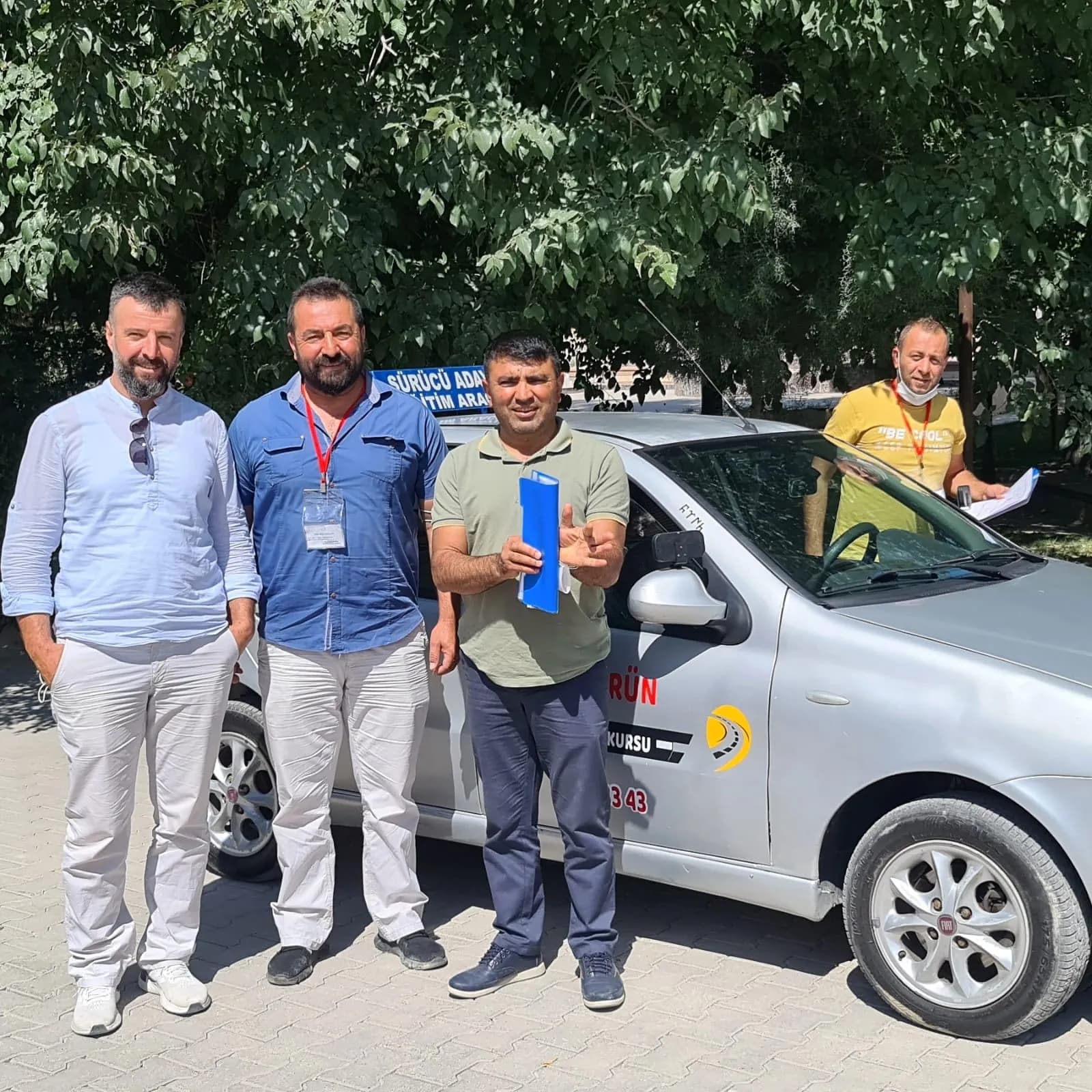 Ağaç gölgesinde gümüş renkli eğitim aracının önünde poz veren dört erkek; ikisi kırmızı askılı yaka kartı taşıyor