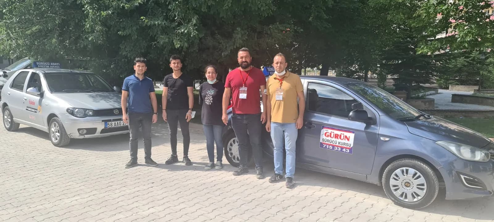 İki eğitim aracının önünde yan yana duran dört erkek ve bir kadın; ikisi boynunda kırmızı askılı yaka kartı taşıyor