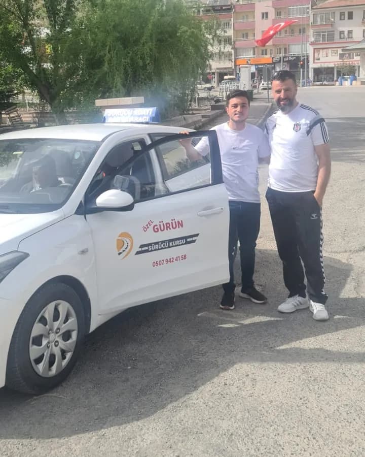 Beyaz eğitim aracının açık kapısı yanında poz veren iki erkek; arkada Türk bayrağı ve dağ eteğindeki yerleşim görünüyor