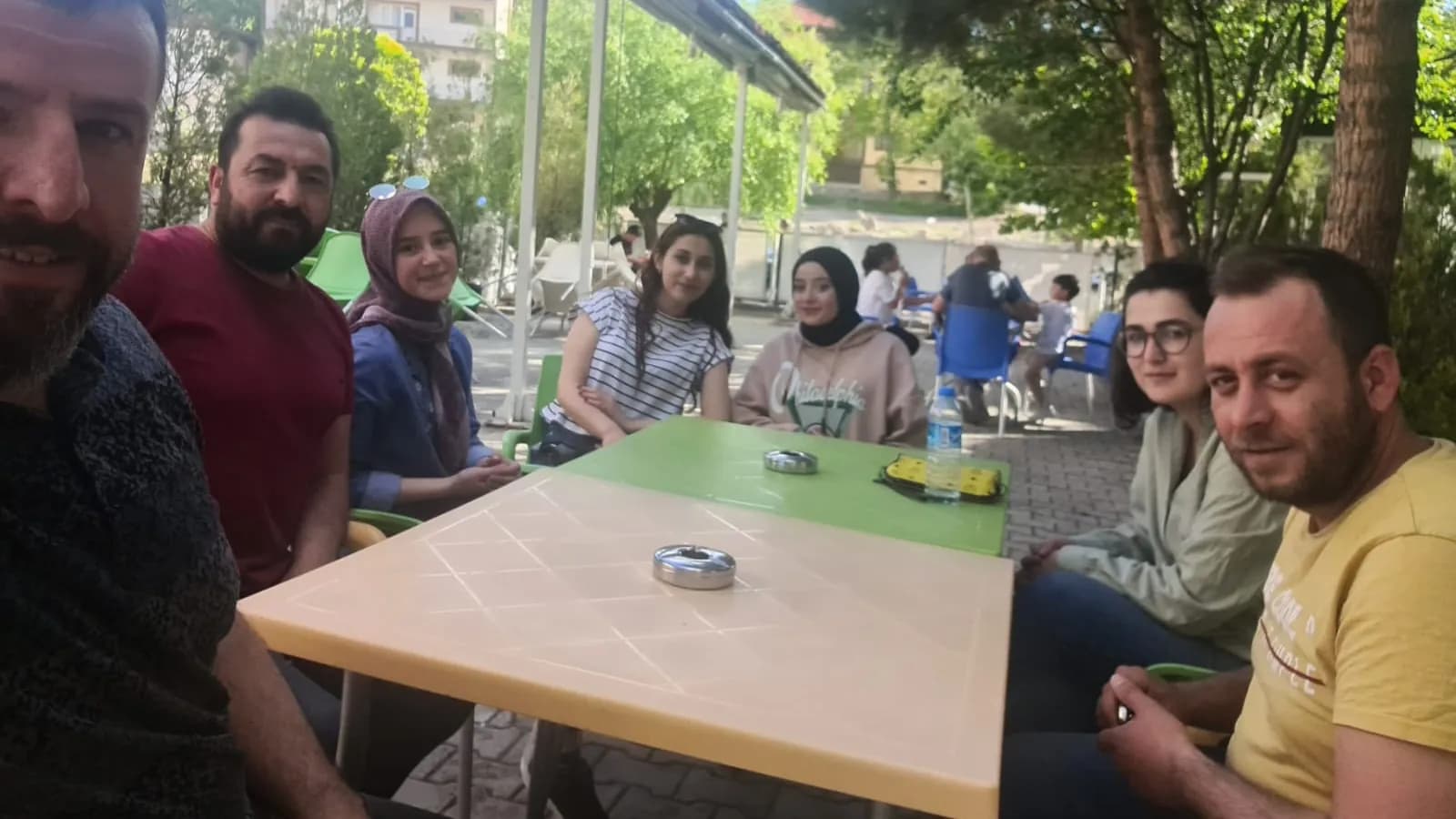 Çay bahçesinde masa etrafında oturup kameraya bakan üç erkek ve dört kadın; eğitim arasında verilmiş bir mola