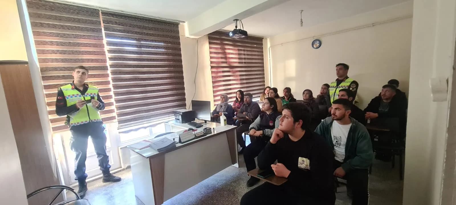 Sınıfta yansıtıcı yelekli iki üniformalı görevli ve onları dinleyen kursiyerler; tavanda projeksiyon cihazı asılı