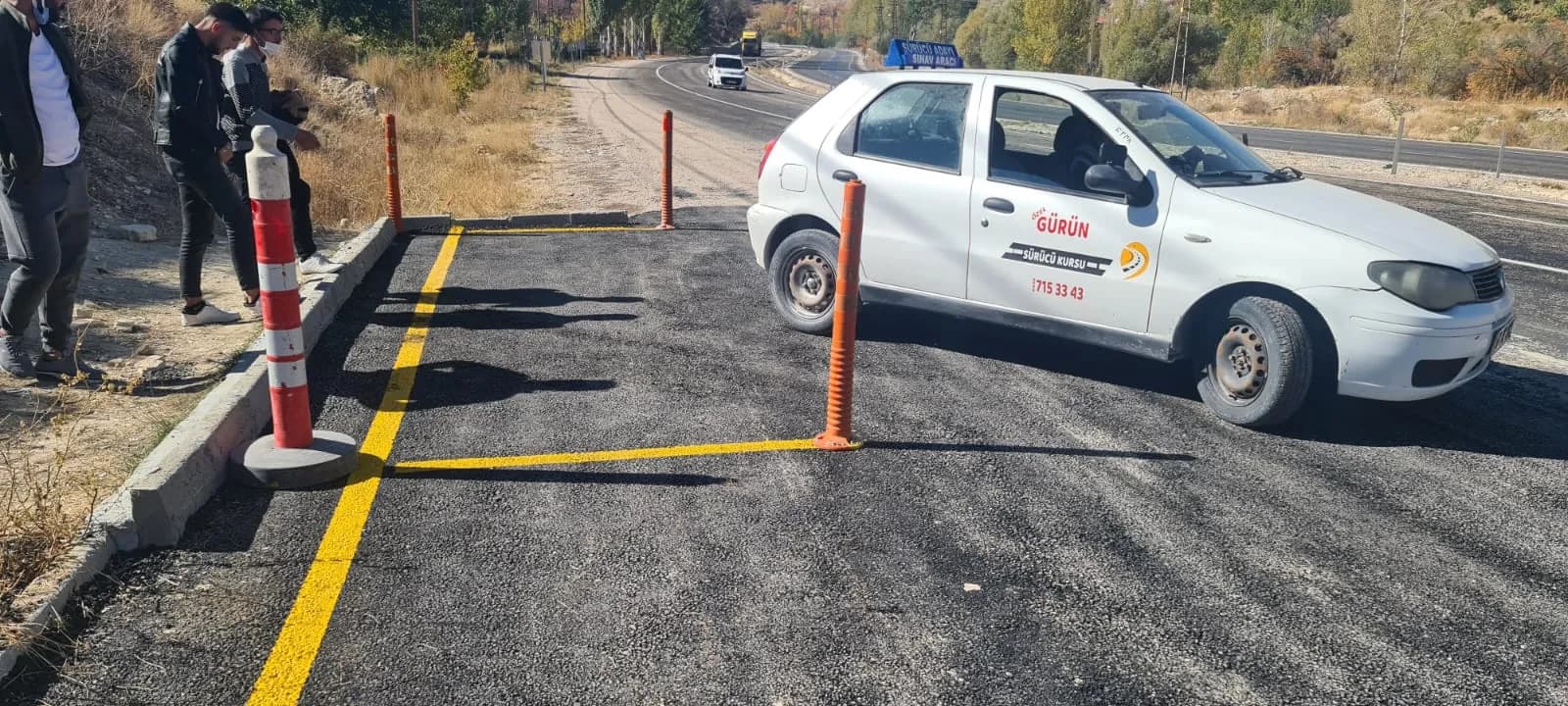 Yeni asfaltlanmış direksiyon sahasında sarı çizgili park alanı, turuncu dubalar ve manevra yapan eğitim aracı