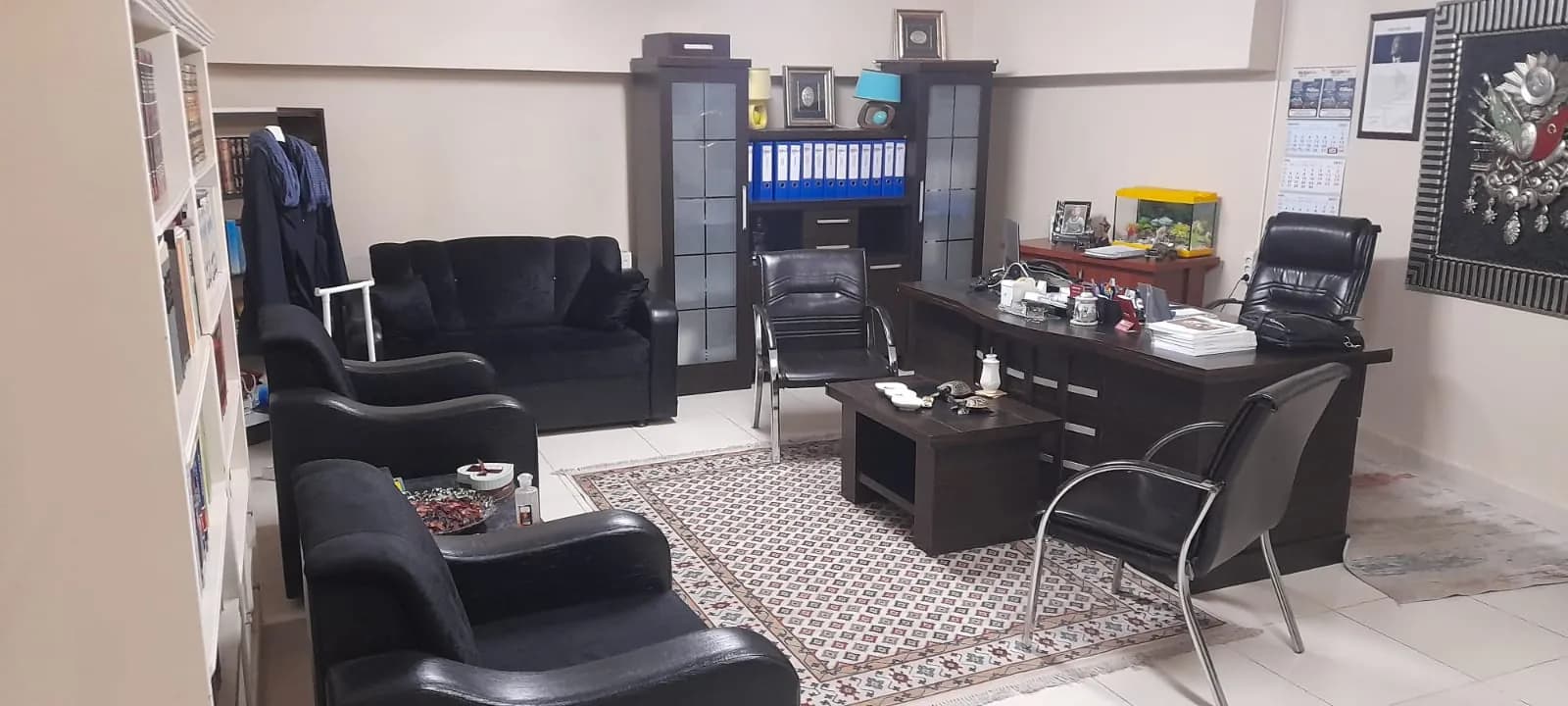 Siyah deri koltuk takımı, çalışma masası, dosya dolabı ve kitaplığı olan ofis odası; zeminde desenli kilim var